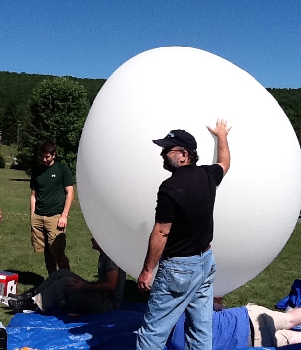 John Moore stands beside a High Altitude Balloon to prepare for a launch; other people are sitting or standing around the balloon helping prepare
