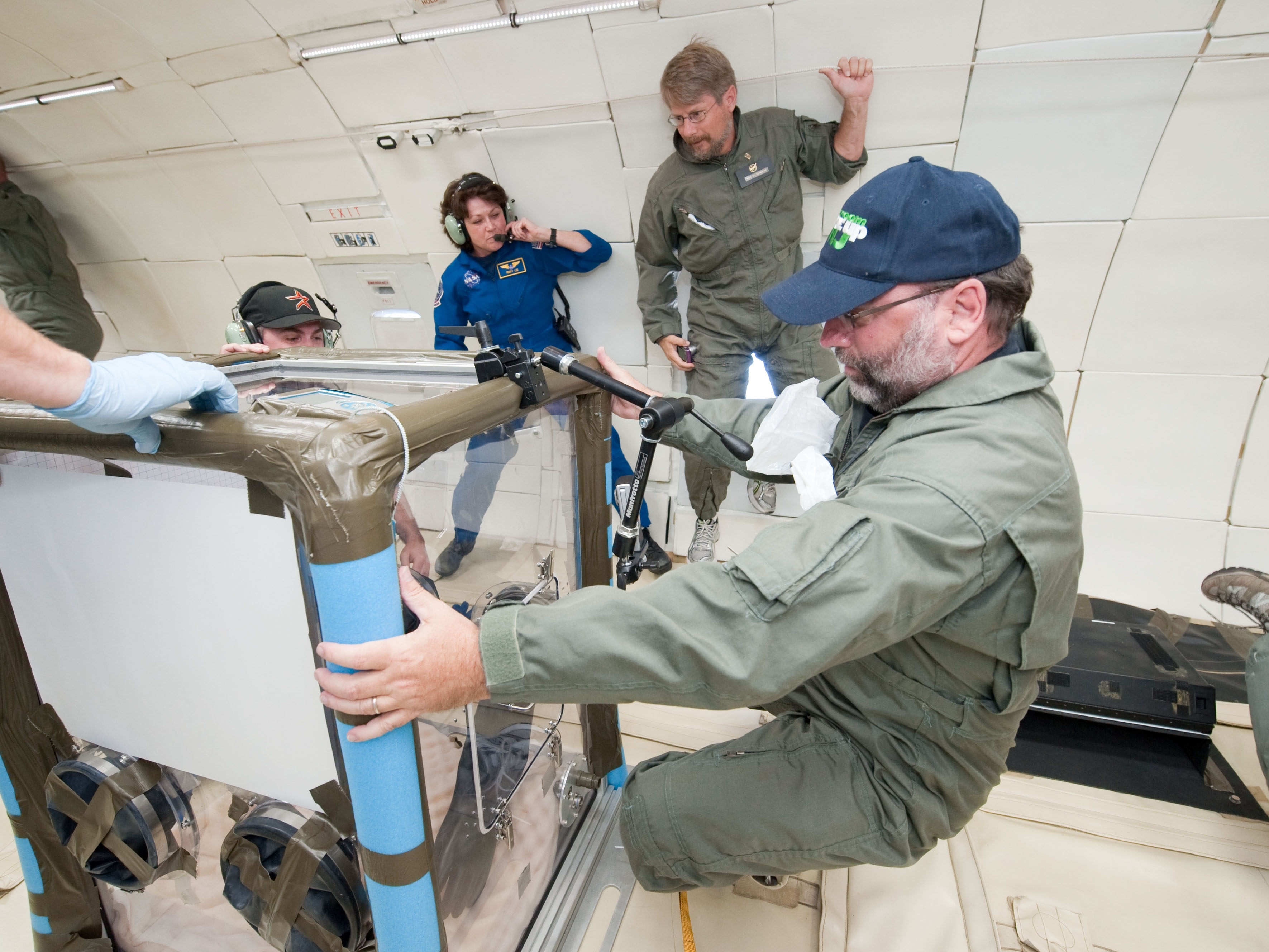 John Moore maneuvers a CubeSat (small satellite) onboard a NASA zero-gravity aircraft; the CubeSat appears to be about four-feet square. there are other people in the aircraft behind the cubesat