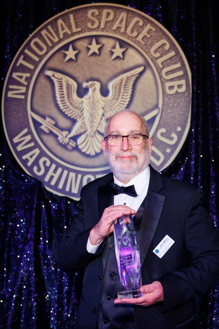 Wearing a tuxedo, John Moore stands in front of a placard that reads National Space Club Washington D.C. and holds a crystal trophy